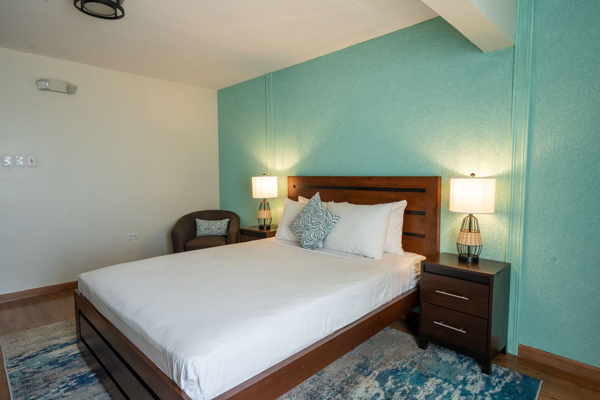 A neatly made bed with white linens and blue accent pillows is centered in a room with teal walls. Two side tables with lamps are on either side, and there is a chair in the corner.