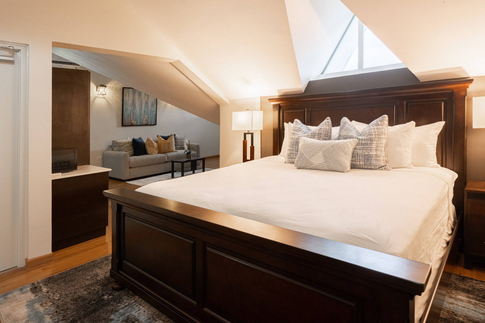 A bedroom with a large wooden bed, white bedding, and decorative pillows. A sitting area with a sofa and cushions is visible under a sloped ceiling. A window allows natural light into the room.