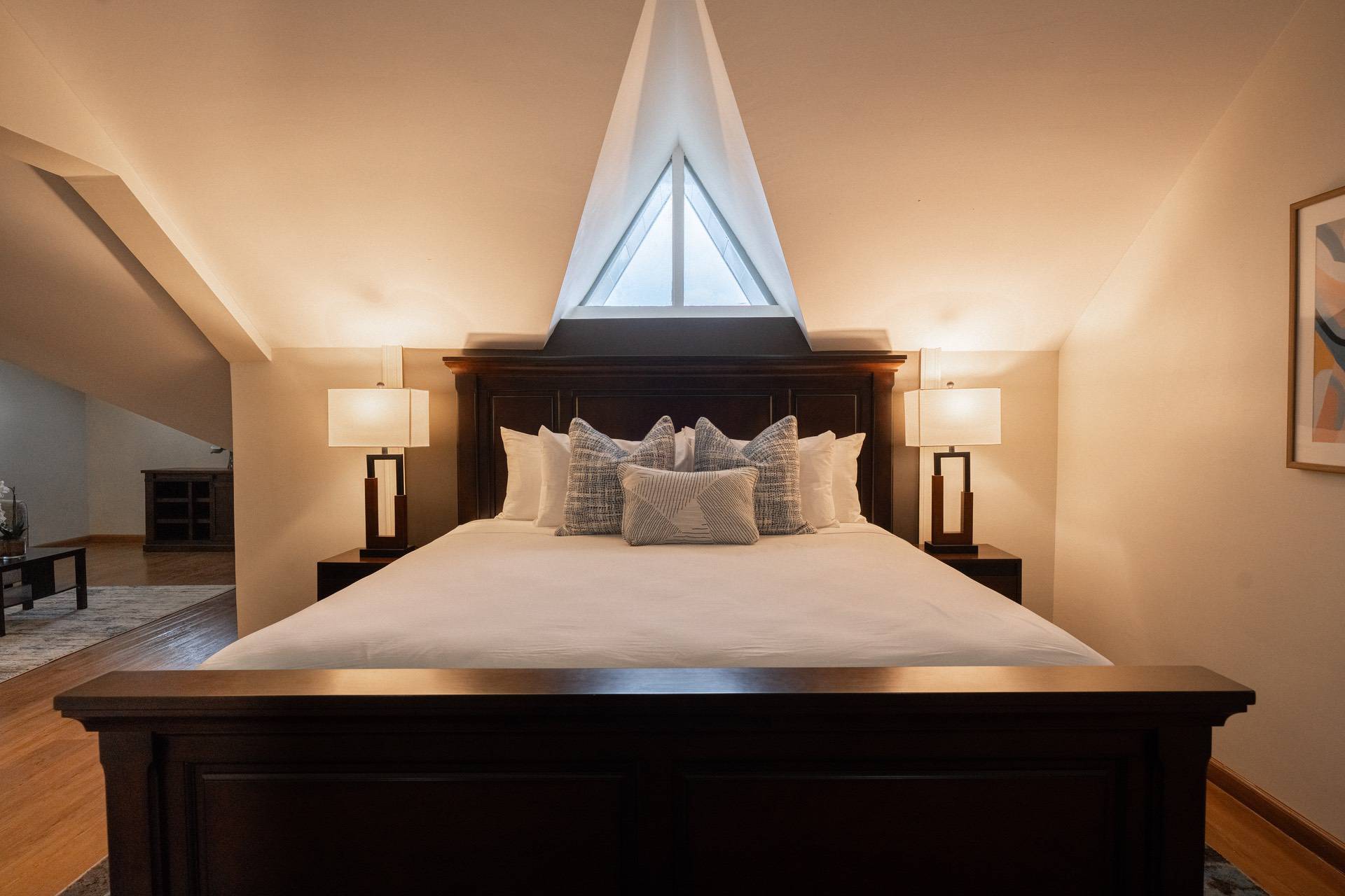 A bedroom with a large wooden bed, white linens, and multiple pillows. Two small lamps sit on each side of the bed, and there is a triangular window above the headboard.