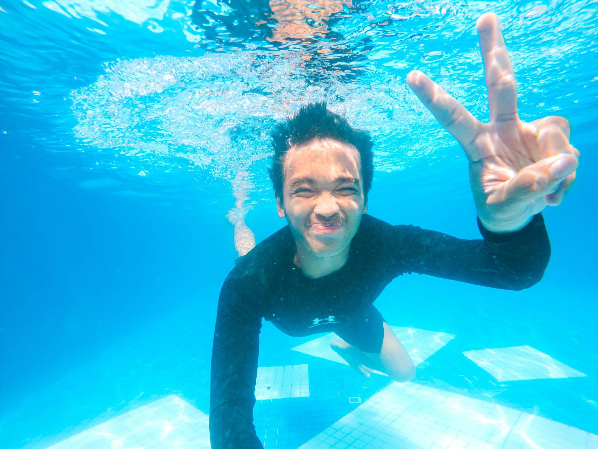 Person in a swimming pool underwater, wearing a black swim shirt, with a playful facial expression, and making a peace sign with one hand.