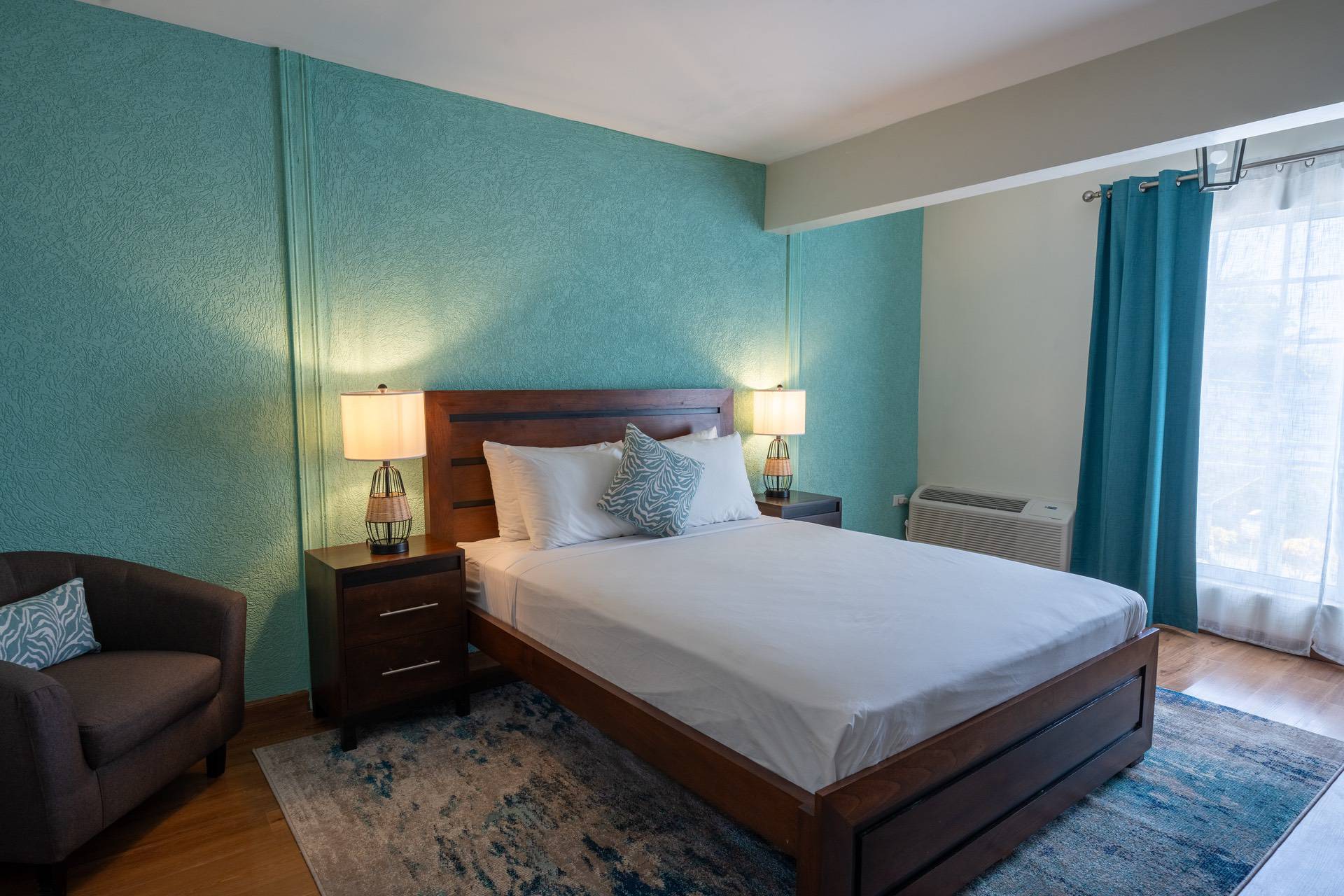 A neatly made queen-sized bed with white linens, wooden frame, flanked by two nightstands with lamps in a room with teal walls, a gray armchair, and blue curtains.